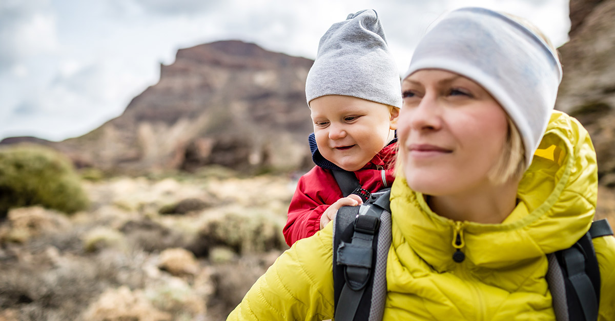 mother wearing a yellow jacket and white hat carrying a baby wearing a red jacket in a backpack
