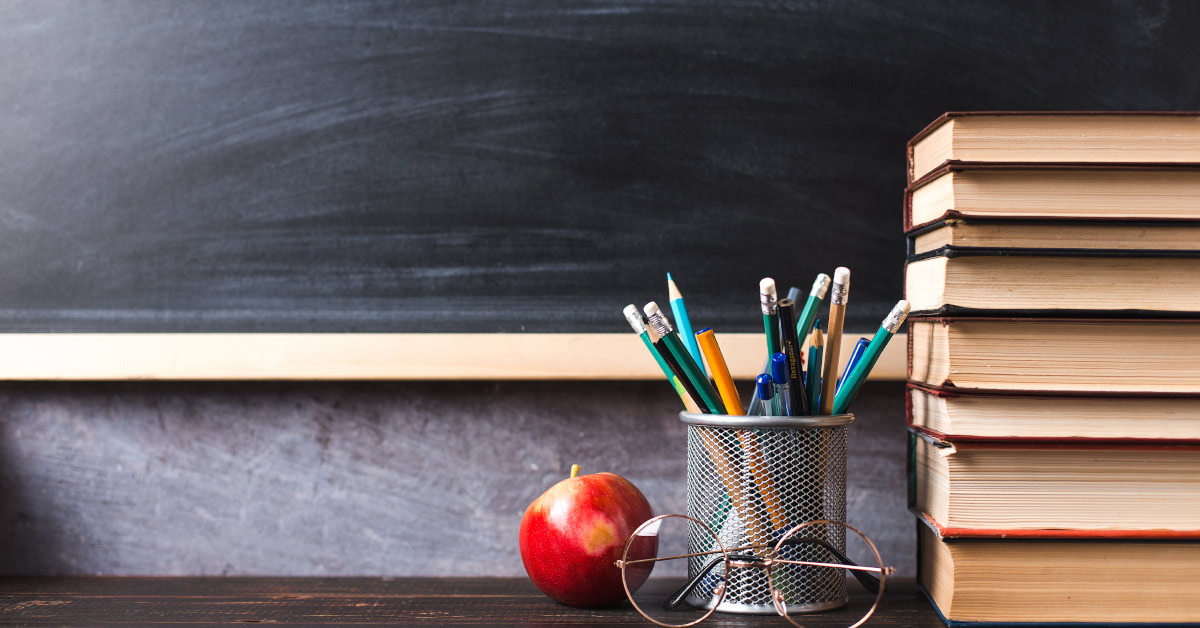 teachers desk with stack of books pencils and red apple. a blackboard can be seen in the background