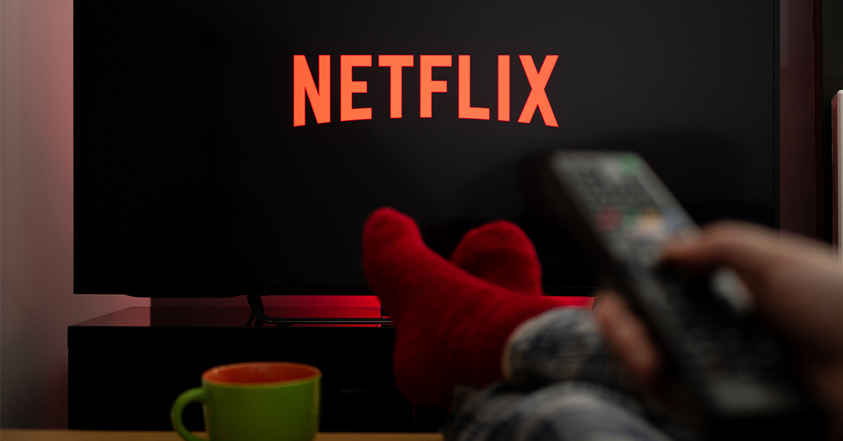 Netflix on TV screen. Person sitting with legs crossed and remote pointed at television.