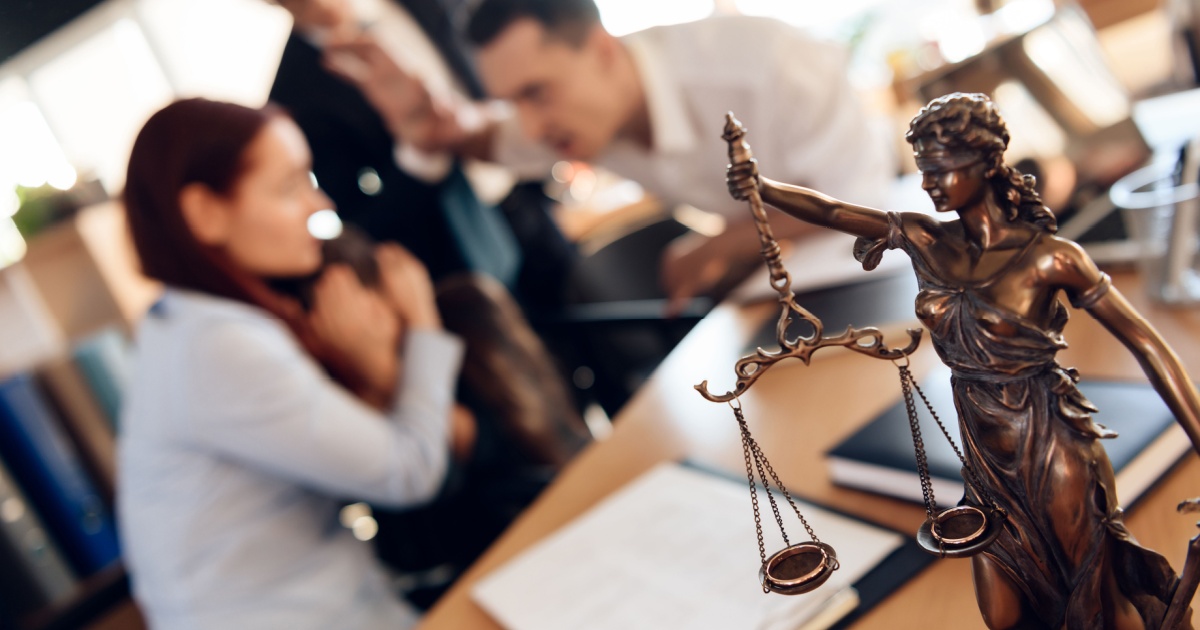 Bronze figurine of Themis holds wedding rings on scales of balance, being in foreground. Couple divorce. Adult couple gets divorced from attorney for divorce in office.