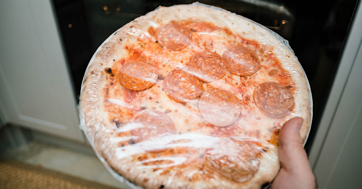 person holding a frozen pepperoni pizza that is still in its plastic wrapping