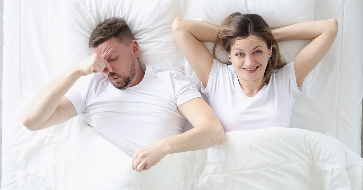 couple wearing white laying in bed. Woman is smiling with arms behind her head while man cowers over pinching nose from foul smell. Couples farting in front of each other