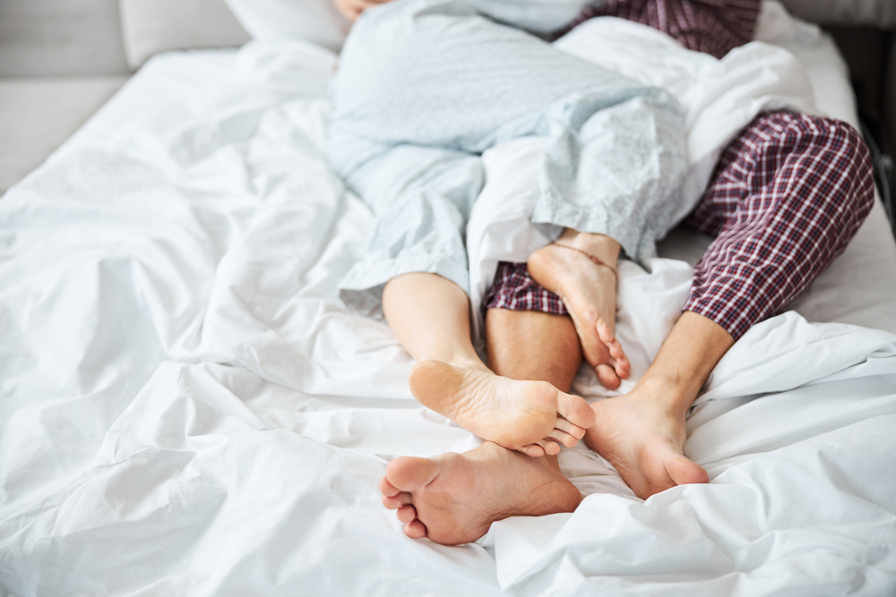 Close up of barefoot man in pajamas cuddling with girlfriend while lying on white sheets