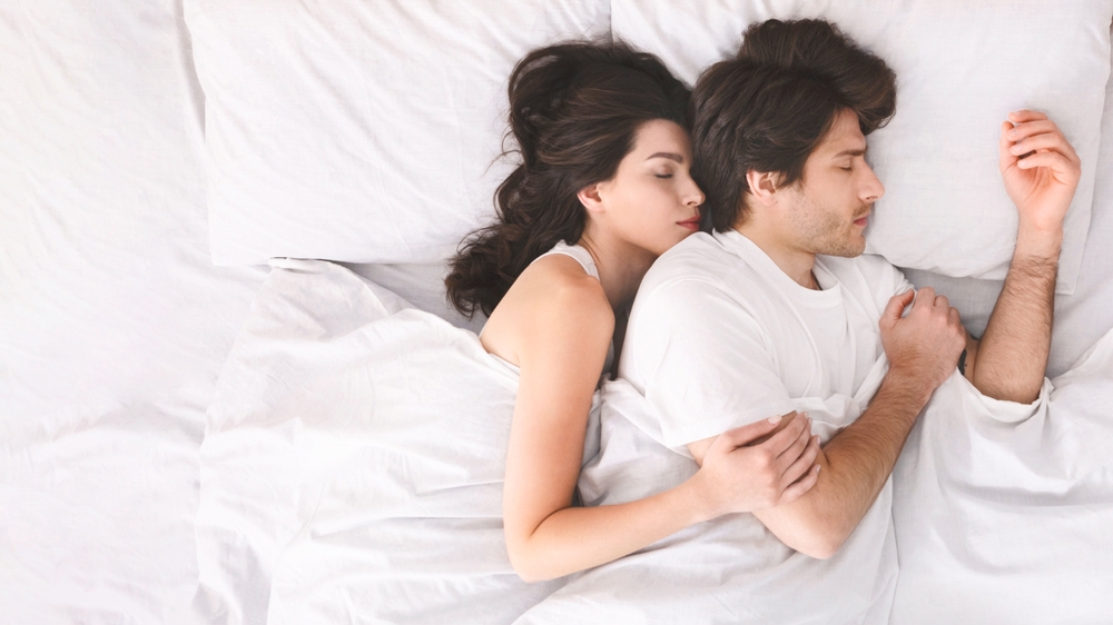 A couple is sleeping peacefully in bed together, with the woman nestled against the mans back, wearing white shirts and are completely relaxed, suggesting a comfortable and loving relationship.