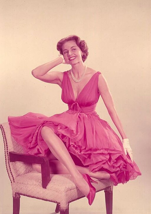 Cyd Charisse poses elegantly in a rose-colored dress, her hand gracefully positioned near her face, embodying the Hollywood glamour that made her a star in the 1950s.