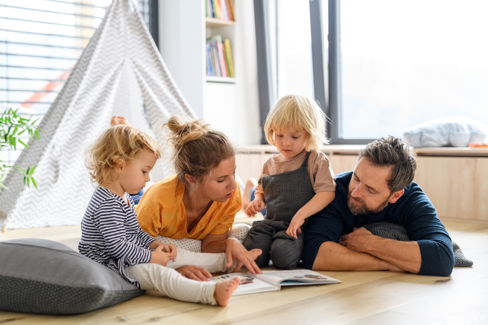 Young nuclear family playing with toys in a living room. Parents and children lying on floor, looking at children's story book, spending weekend day indoors.
